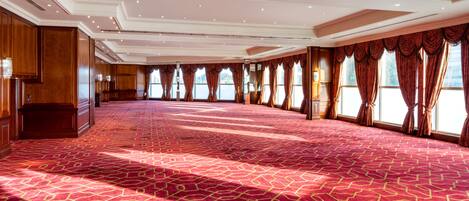 A large, well-lit room with a patterned carpet, wooden paneling, and large windows with curtains.