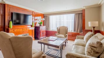 A hotel room with a flat-screen TV, a wooden cabinet, a red decorative piece, a beige armchair, a white sofa, and a lamp.