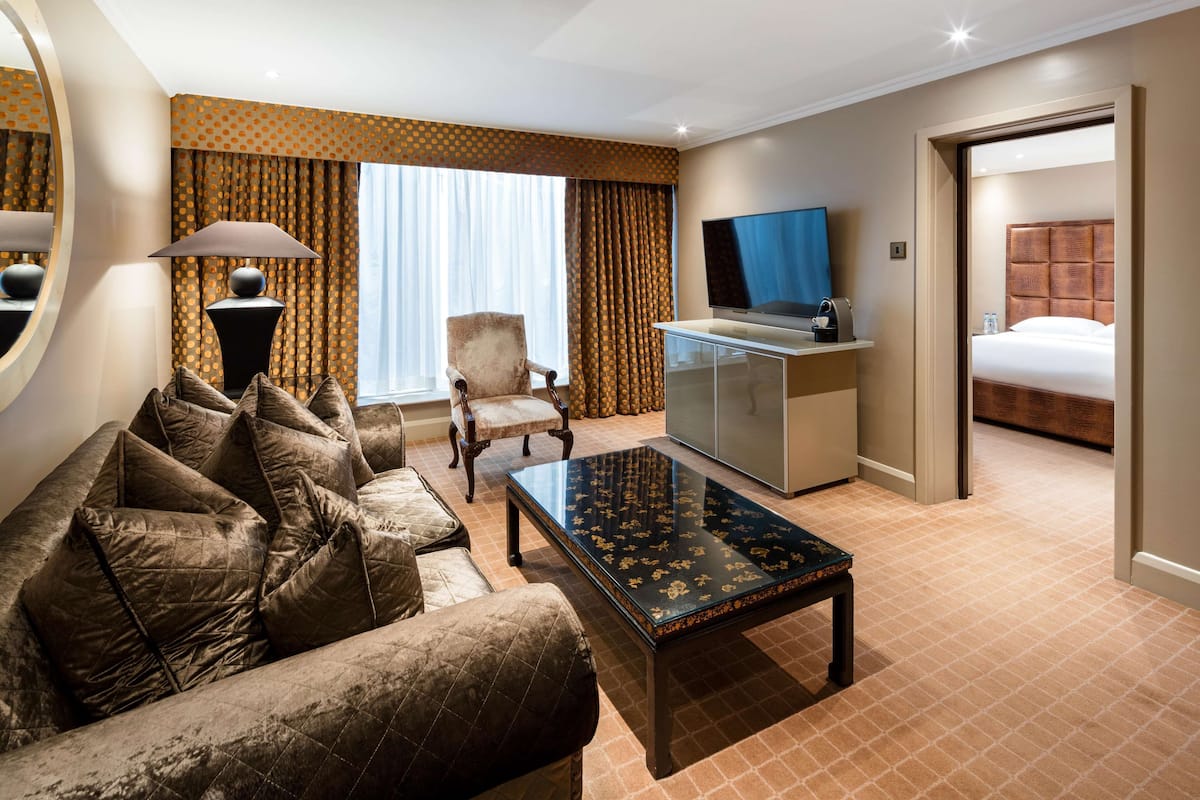 A hotel room with a sofa, armchair, TV, minibar, and a bed.