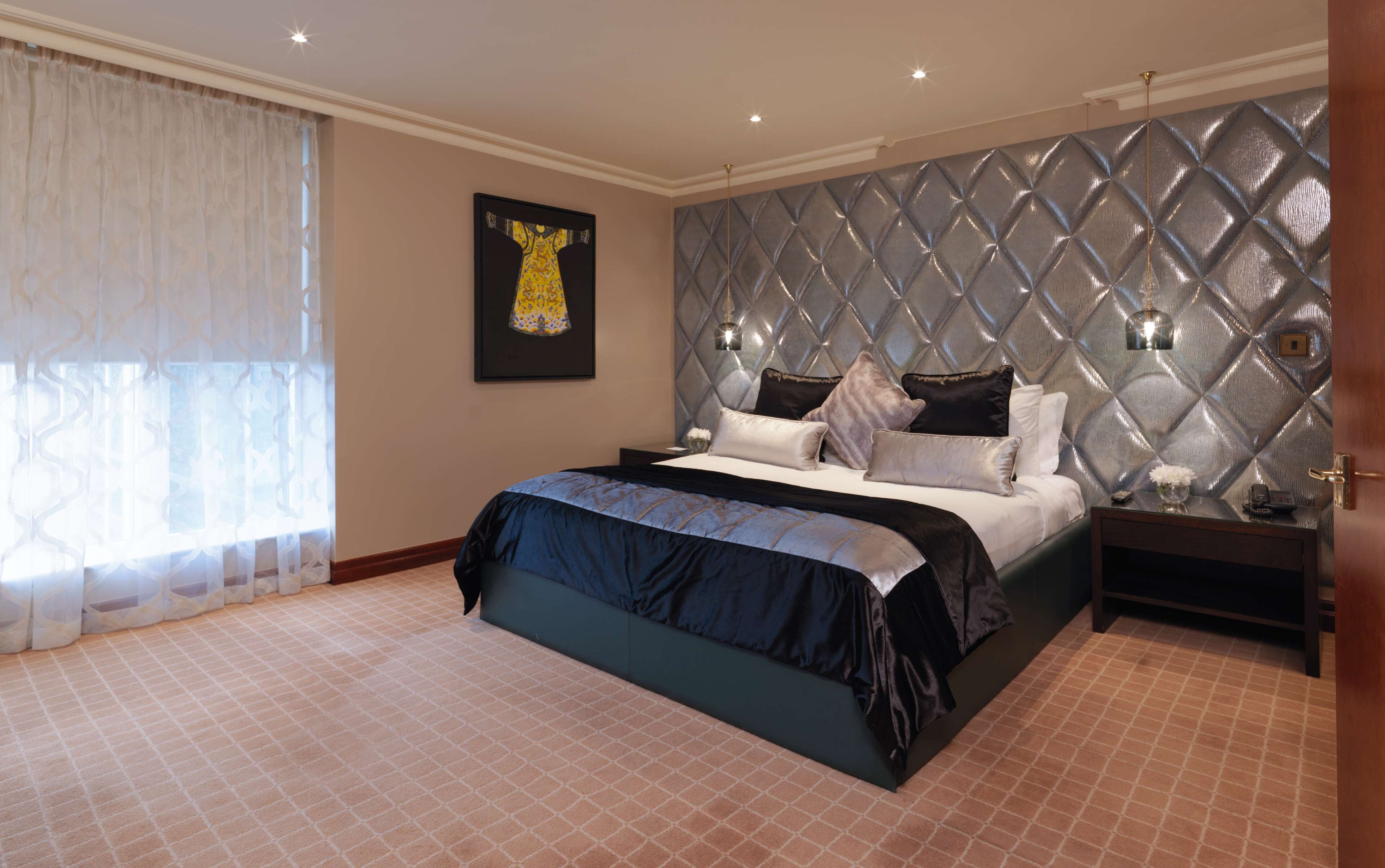 A bedroom with a large bed, a headboard with a diamond pattern, a nightstand with a lamp, and a framed picture on the wall.