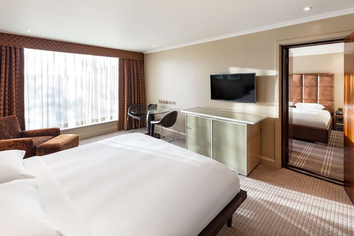 A hotel room with a large bed, a desk with a flat-screen TV, a chair, and a large window with curtains.