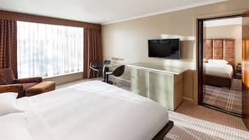 A hotel room with a large bed, a desk with a flat-screen TV, a chair, and a large window with curtains.