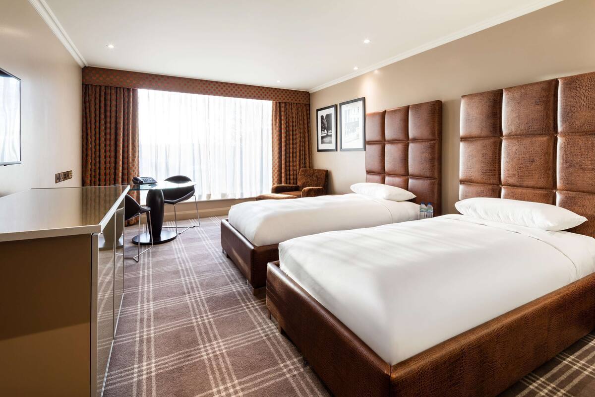 A hotel room with two beds, a desk, a chair, and a large window with curtains.