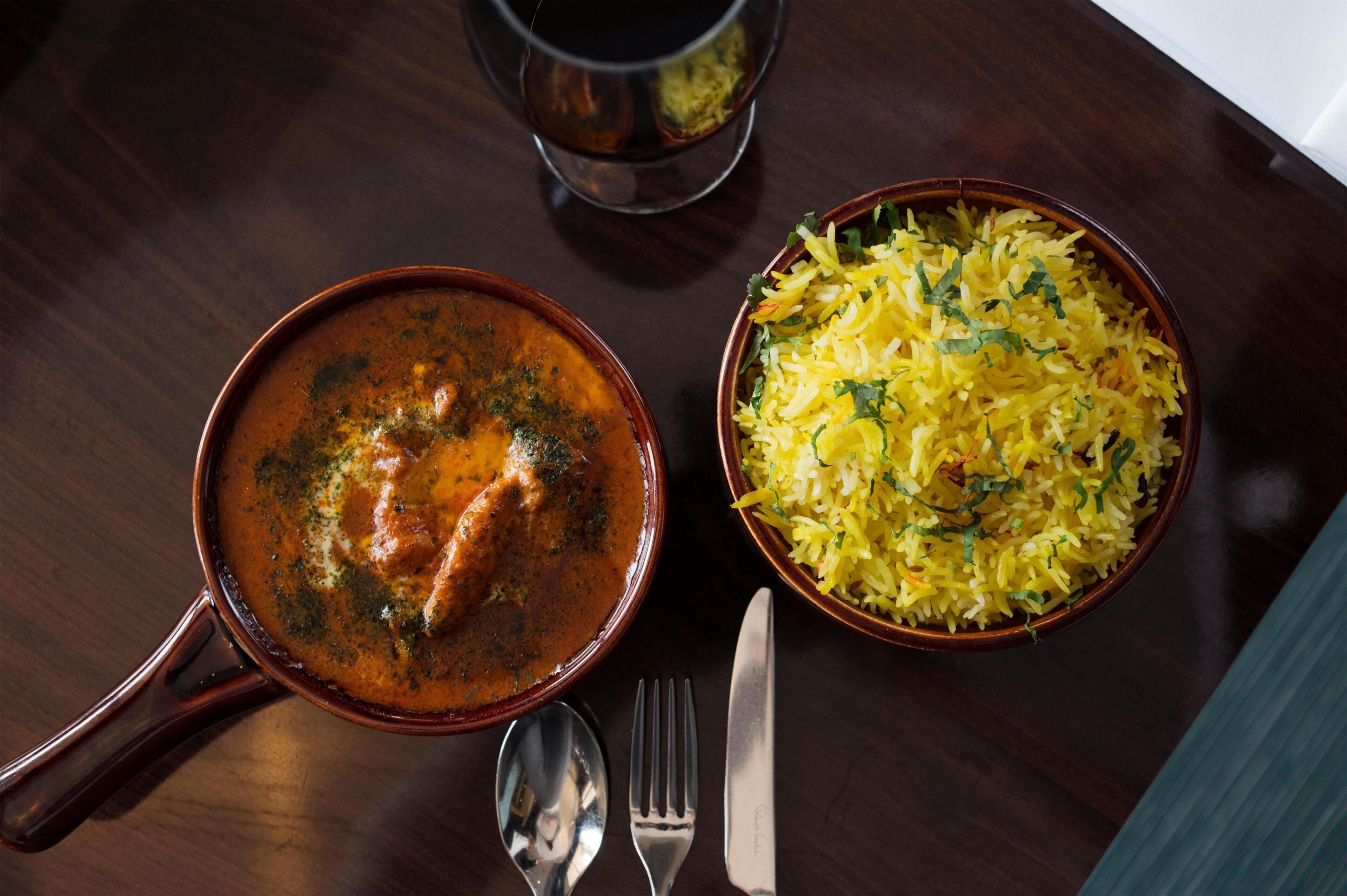 Een tafel met een kom gele rijst, versierd met kruiden, een kom curry, en een glas donkere vloeistof, mogelijk thee of koffie.