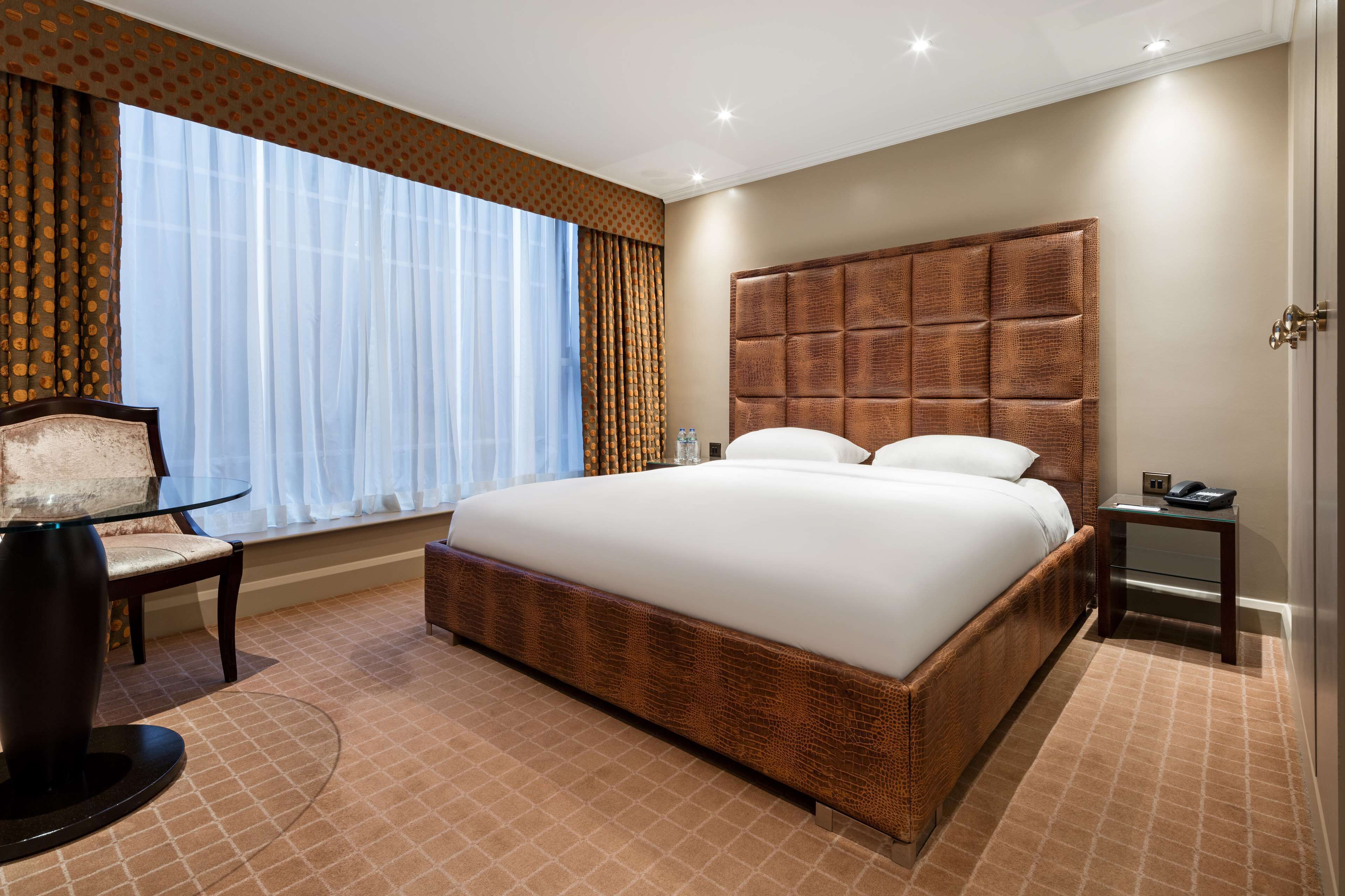 Ein Hotelzimmer mit einem großen Bett, einem Sessel, einem kleinen Tisch und einem Fenster mit Vorhängen.