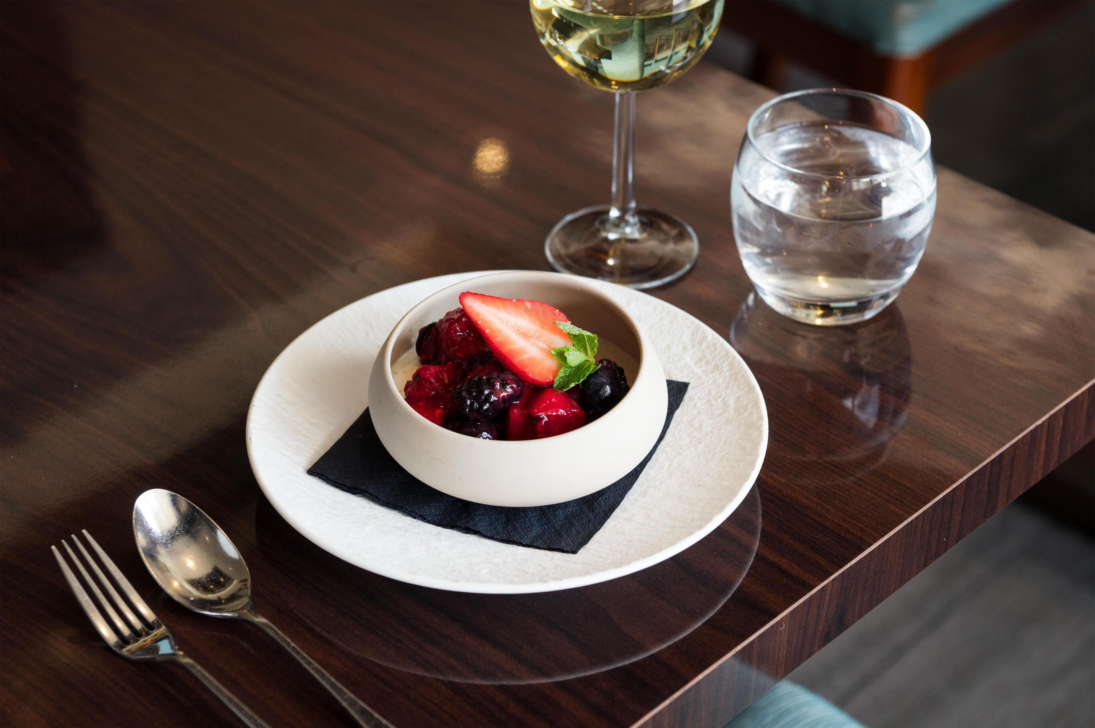A dessert bowl with berries on a white plate, accompanied by a fork and spoon, and a glass of water on a wooden table.