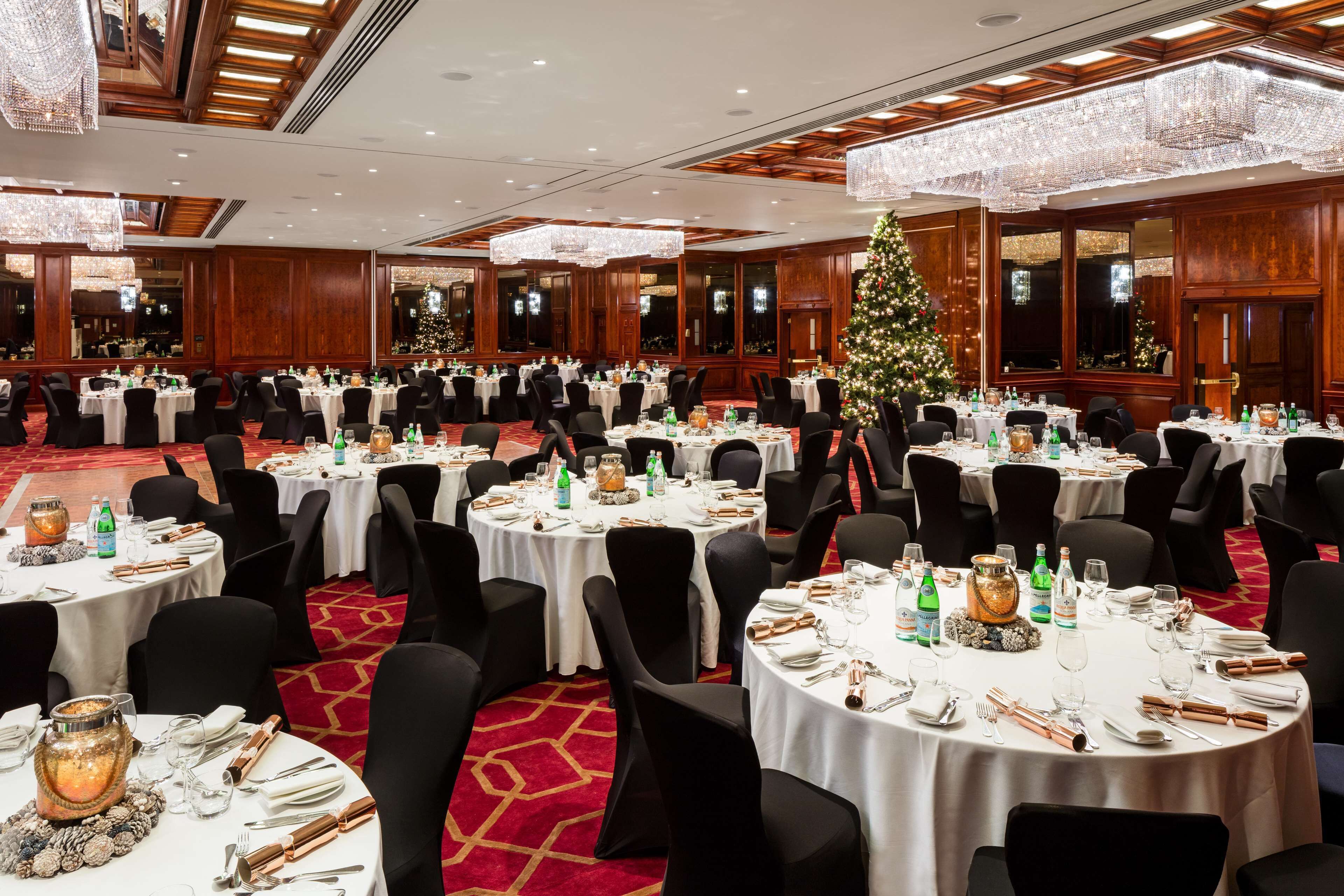 Une salle de banquet avec des tables rondes dressées pour un événement formel, agrémentée de lustres et d’un sapin de Noël.