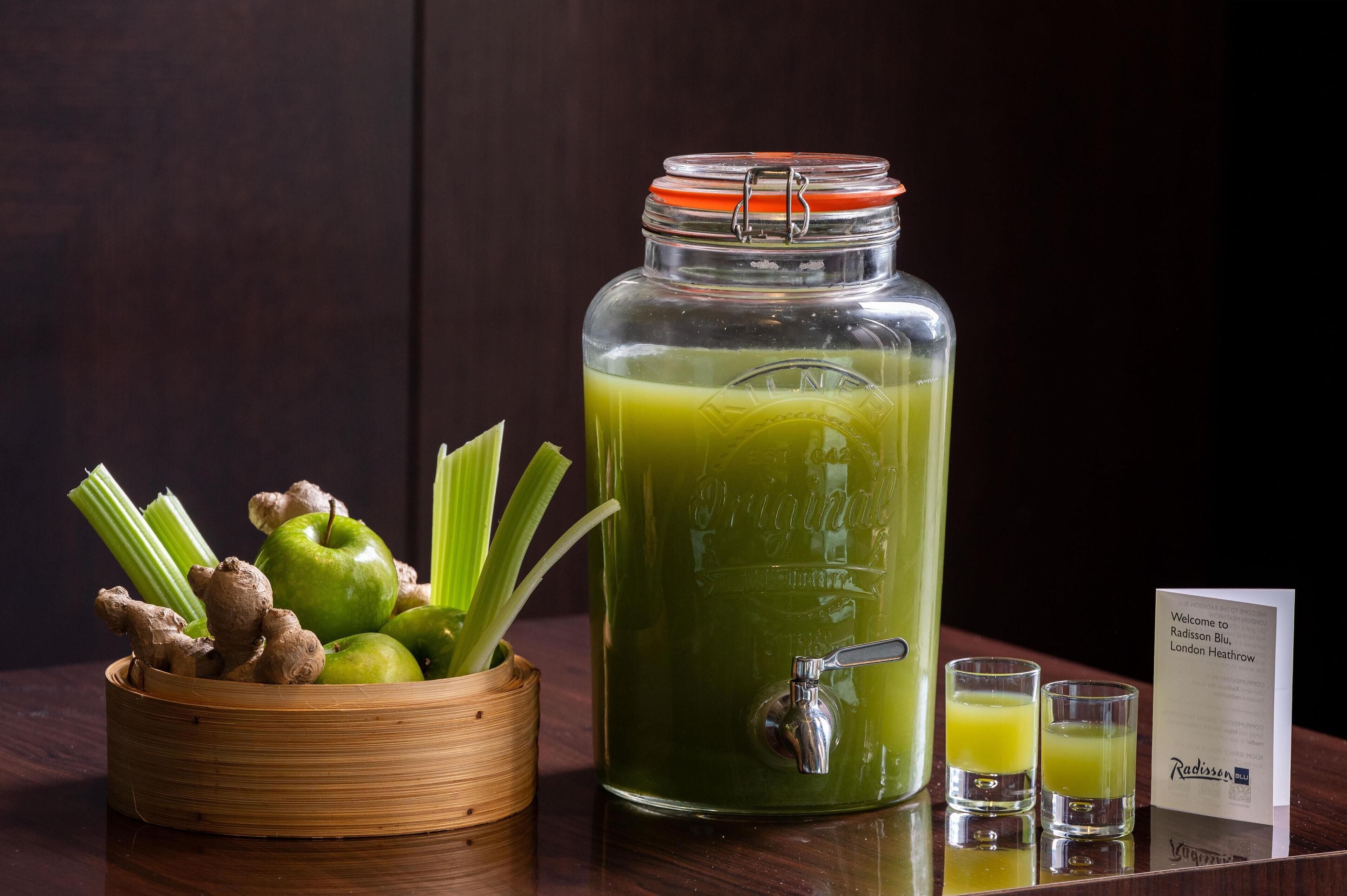 Un frasco grande de vidrio con un grifo, lleno de un líquido verde, y un cuenco de madera con manzanas verdes y apio.