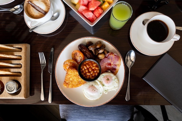 Ein Frühstückstisch mit Eiern, Bohnen, Speck, Pilzen und Toast, dazu eine Tasse Kaffee und ein Glas Saft.