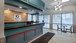 A hotel reception area with a wooden panel wall, a sign reading 'Quality by Choice Hotels,' a reception desk, two chairs, and a large window. - Quality Inn & Suites (Niagara Falls)