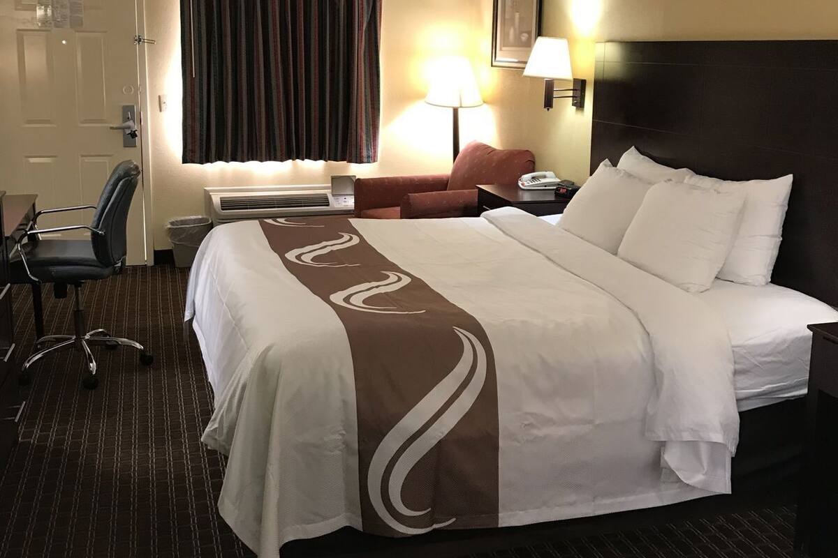 A hotel room with a bed, a desk, a chair, a lamp, and a picture on the wall.