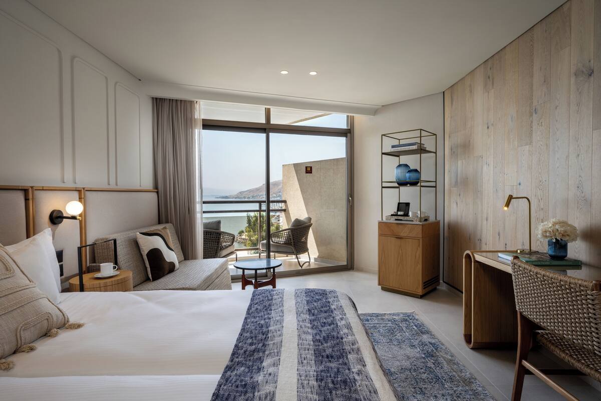 A modern hotel room with a large bed, a seating area with a view, a wooden desk, and a bookshelf.