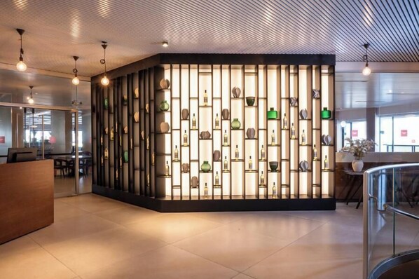 Ett modernt receptionsområde med en central utställning av olika flaskor och vaser, omgivet av en stilren och modern design med ett mönstrat tak och glasräcken.