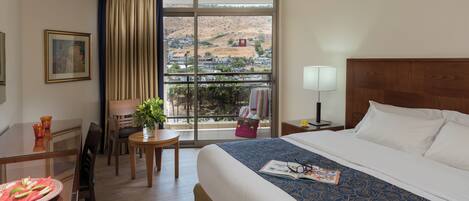 A hotel room with a bed, a desk with a chair, a window with a view of a cityscape, and a lamp.