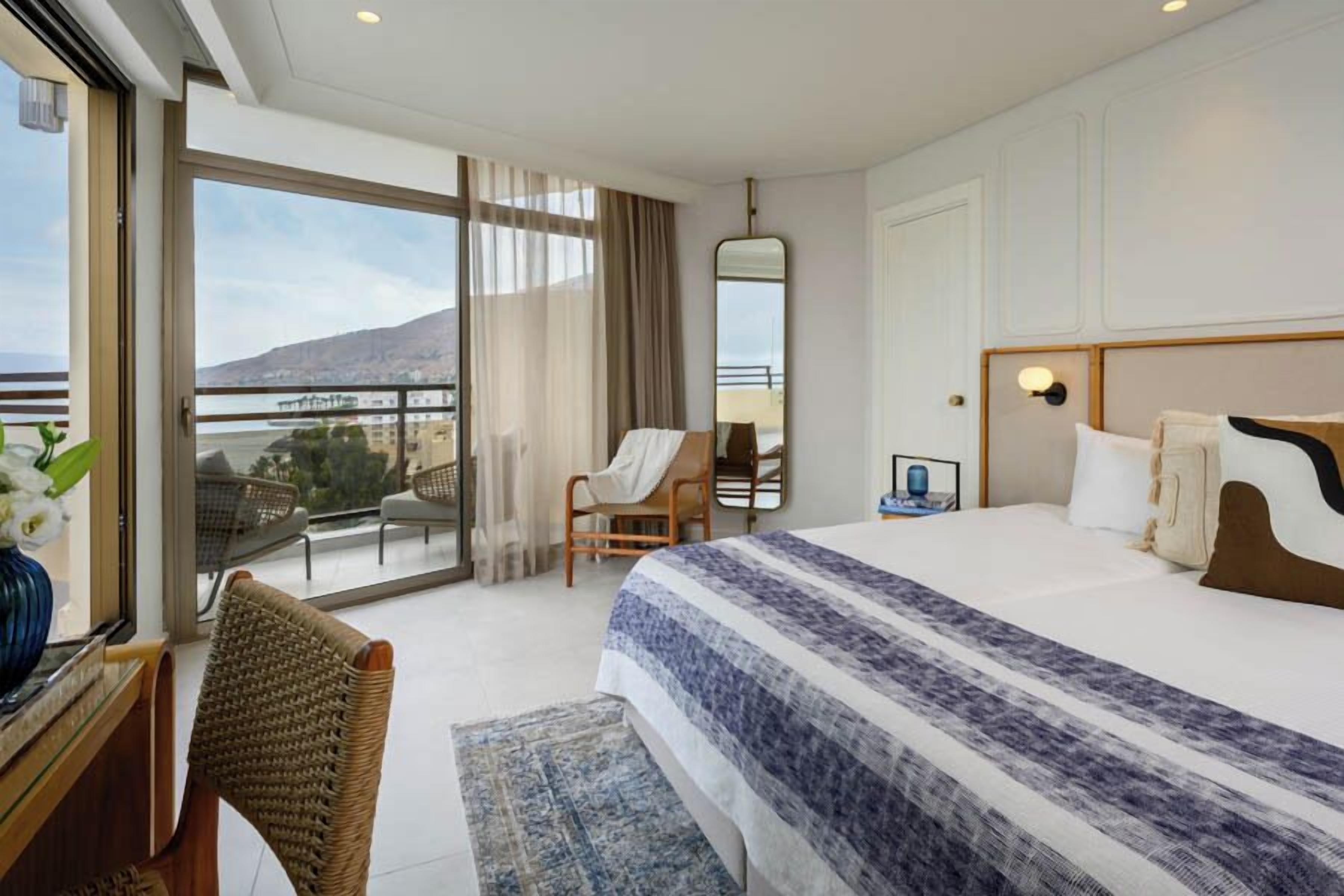 A hotel room with a large bed, a sitting area with a chair, a desk with a vase of flowers, and a view of the sea through a balcony.
