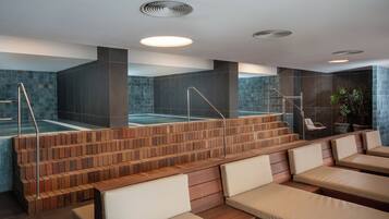 A modern spa area with a wooden staircase leading to a hot tub, cushioned seating, and a wall with a water feature.