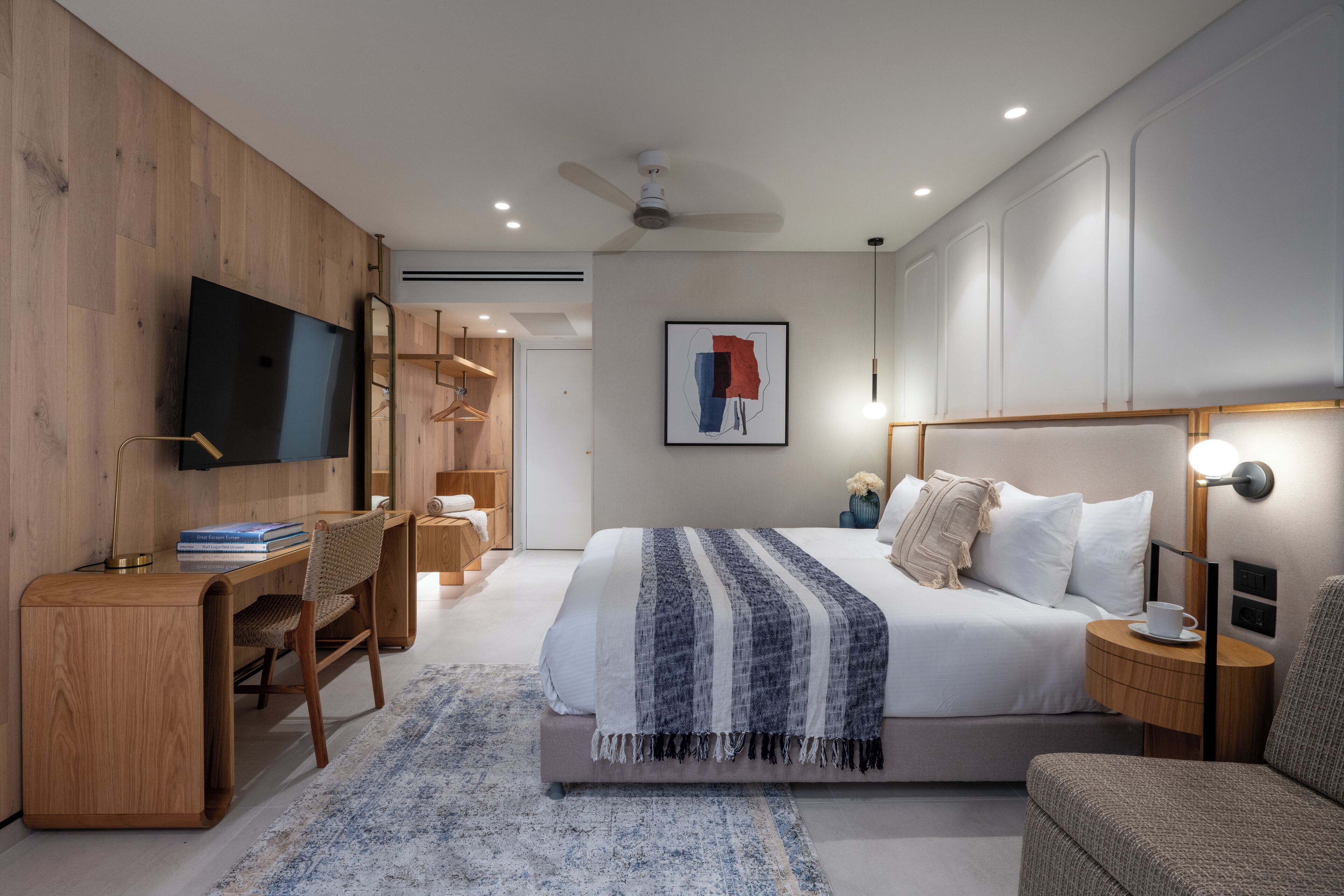 A modern hotel room with a large bed, a desk, a chair, a TV, and a ceiling fan.