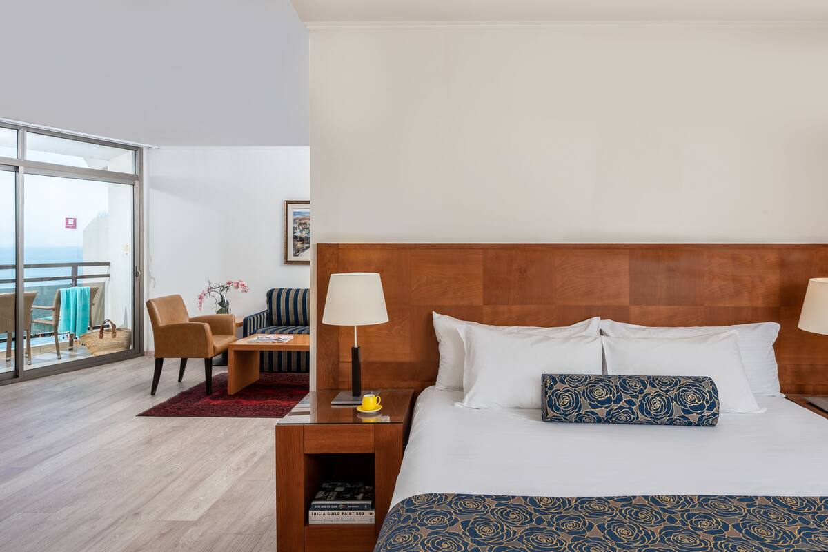 A hotel room with a bed, a nightstand, a chair, a table, a lamp, and a view through a glass door.