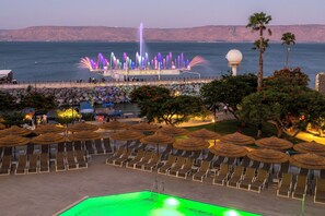 A poolside area with lounge chairs and umbrellas, a water fountain display, and a view of the sea and hills in the background. - Leonardo Plaza Kinneret (Tiberias)