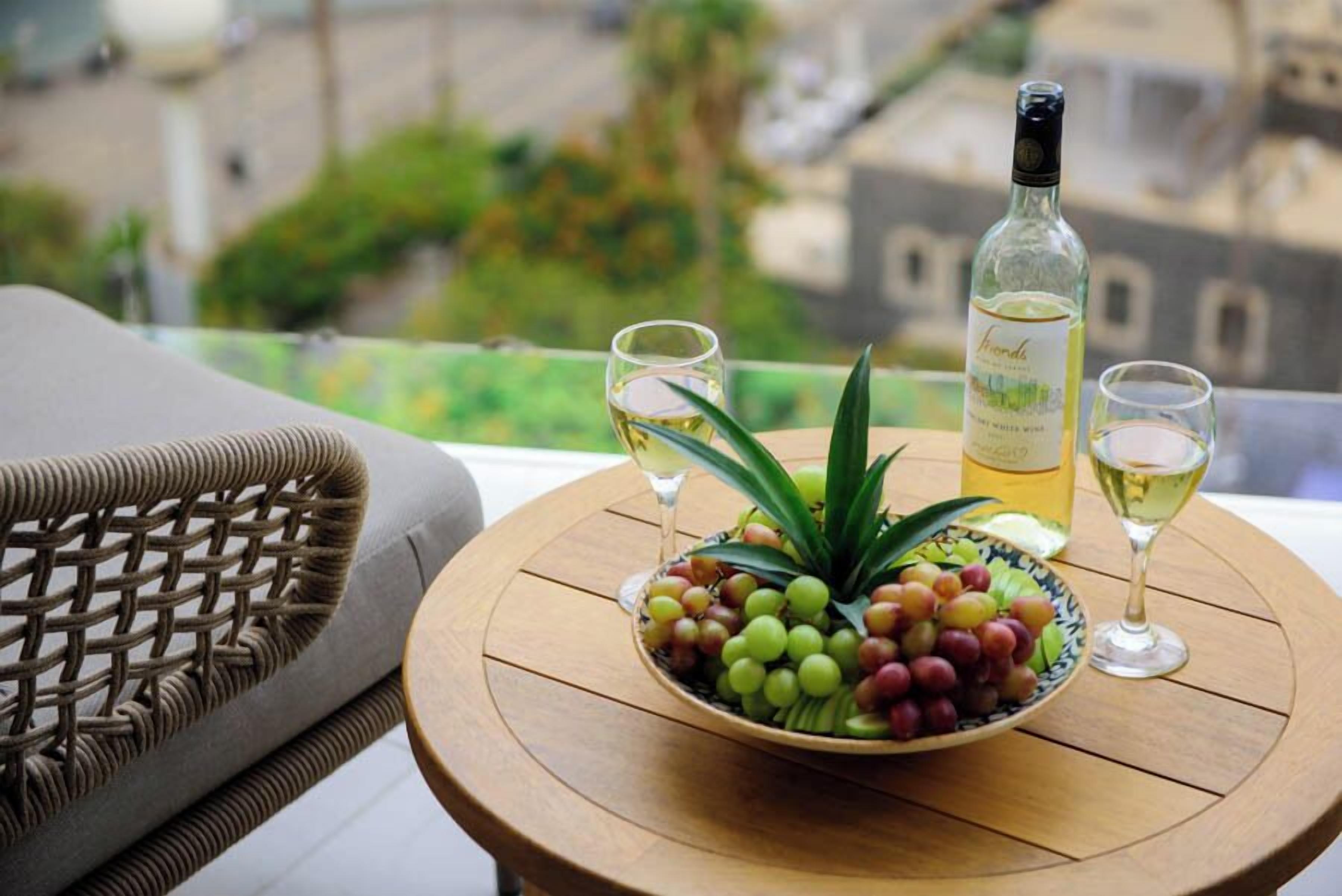 Una silla de mimbre, una mesa redonda de madera con un cuenco de uvas y una botella de vino blanco, junto con dos copas de vino.