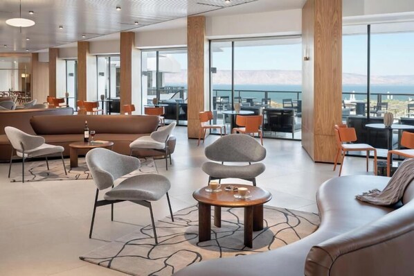 Un vaste espace salon doté de meubles modernes, de grandes fenêtres offrant une vue sur la ville et les montagnes, ainsi qu’un coin bar avec des chaises orange.