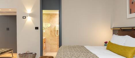 Habitación de hotel con cama, banca, cesta y zona de ducha.