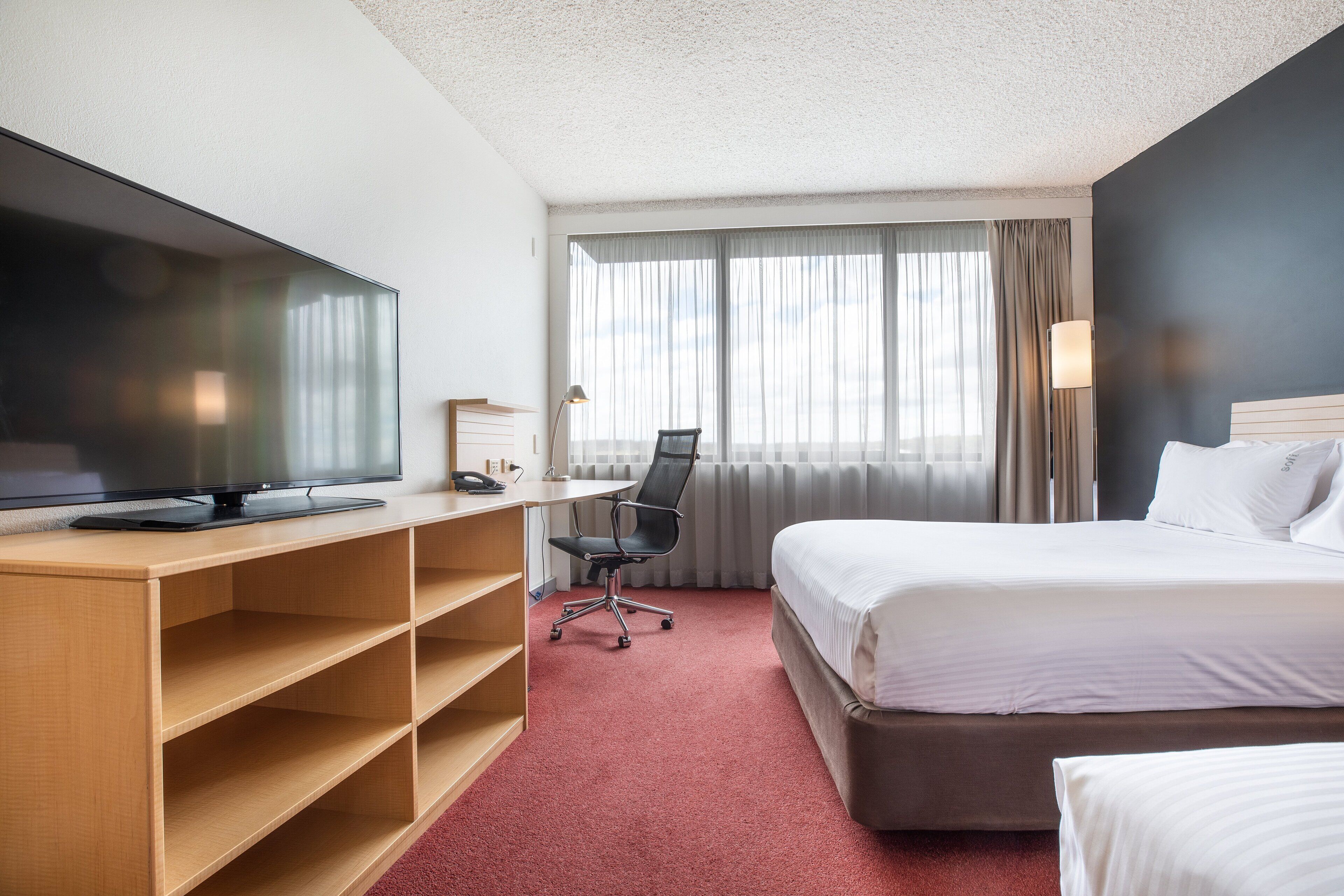 A hotel room with a bed, a desk with a TV, a chair, and a window with curtains.