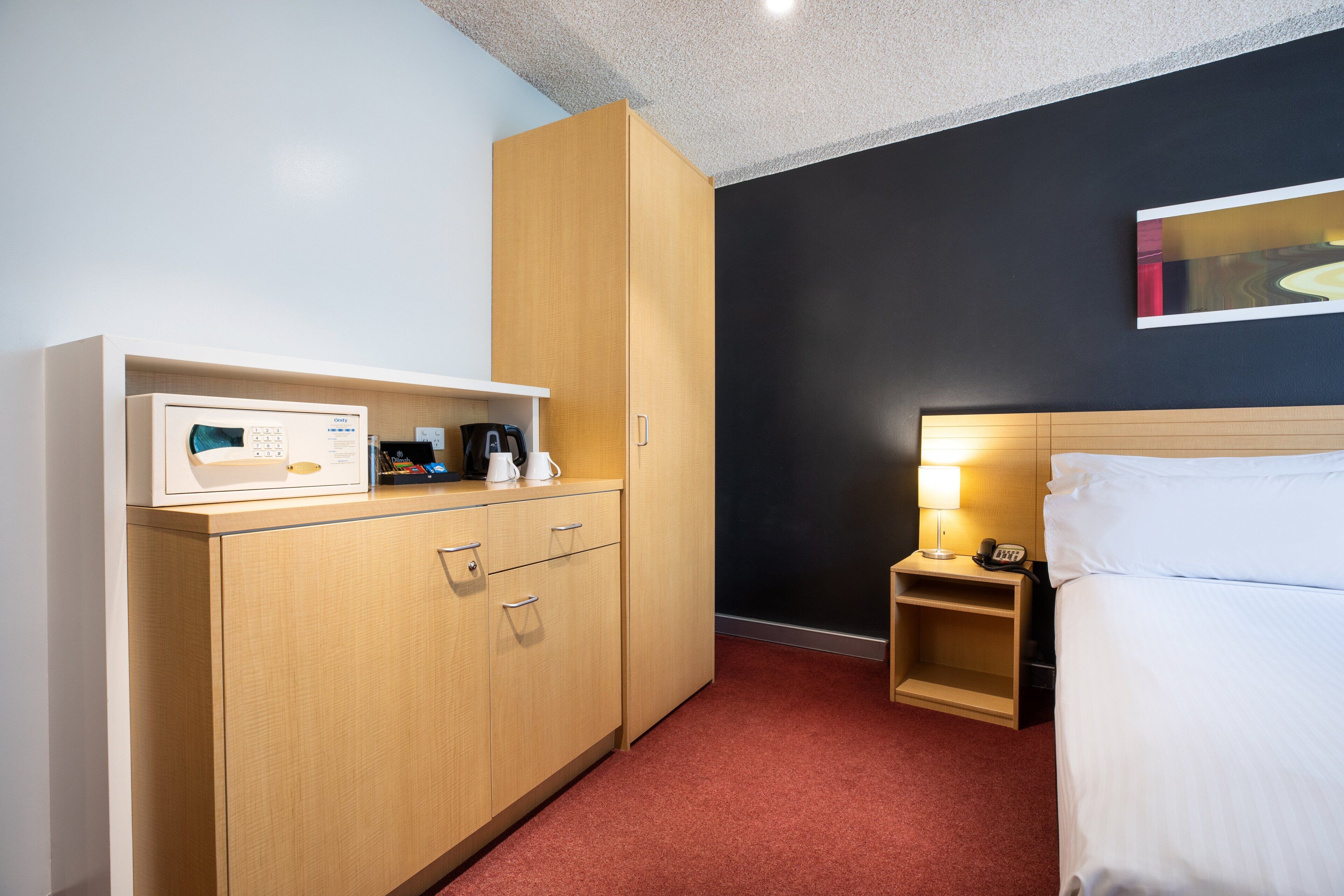 Hotelkamer met een bed, nachtkastje met lamp, een houten garderobe en een klein plankje met een koffiezetapparaat en diverse spullen.