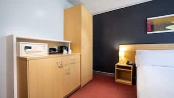 A hotel room with a bed, a nightstand with a lamp, a wooden wardrobe, and a small shelf with a coffee maker and other items.