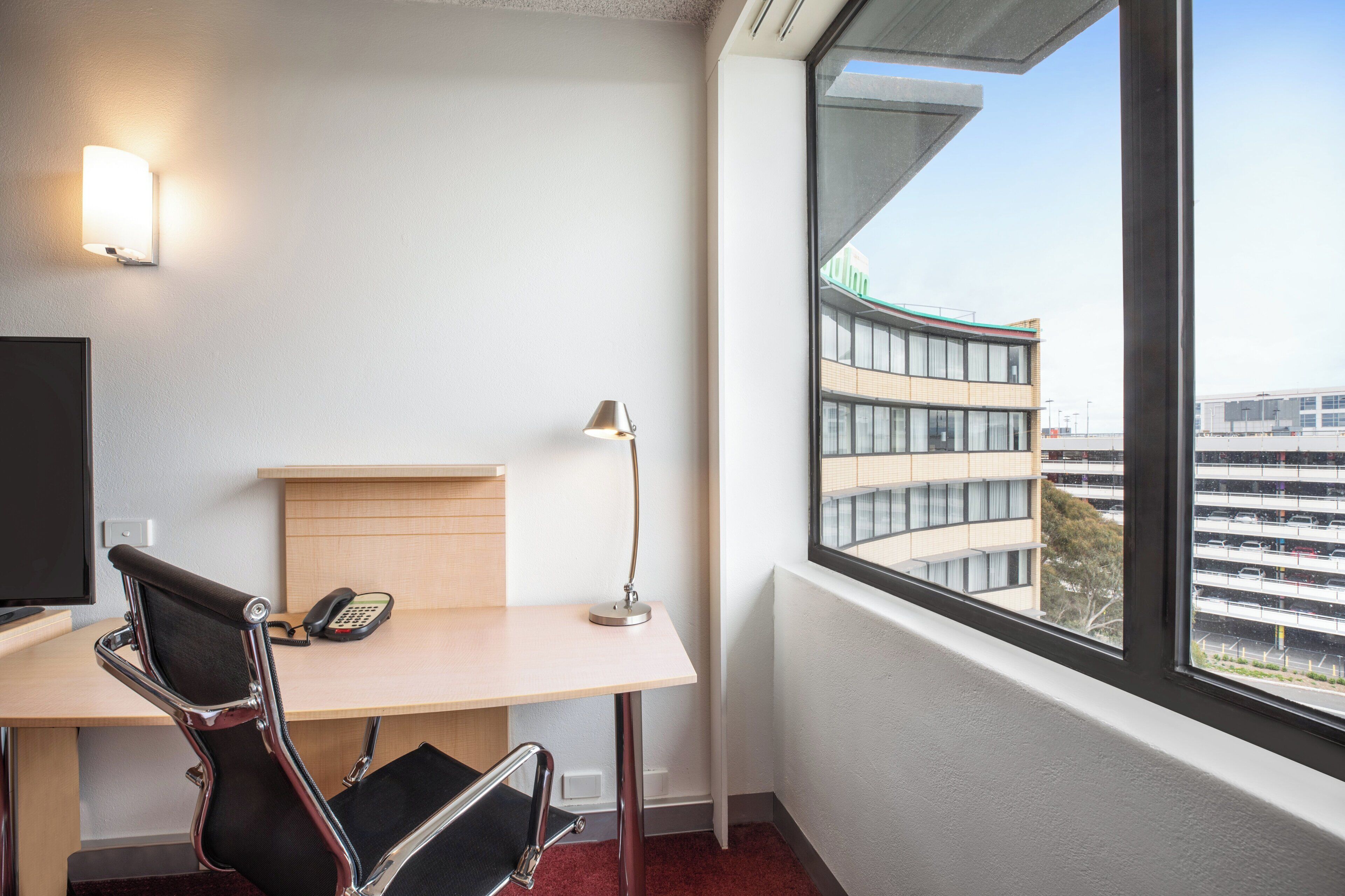 A hotel room with a desk, chair, lamp, phone, and a large window with a view of buildings.