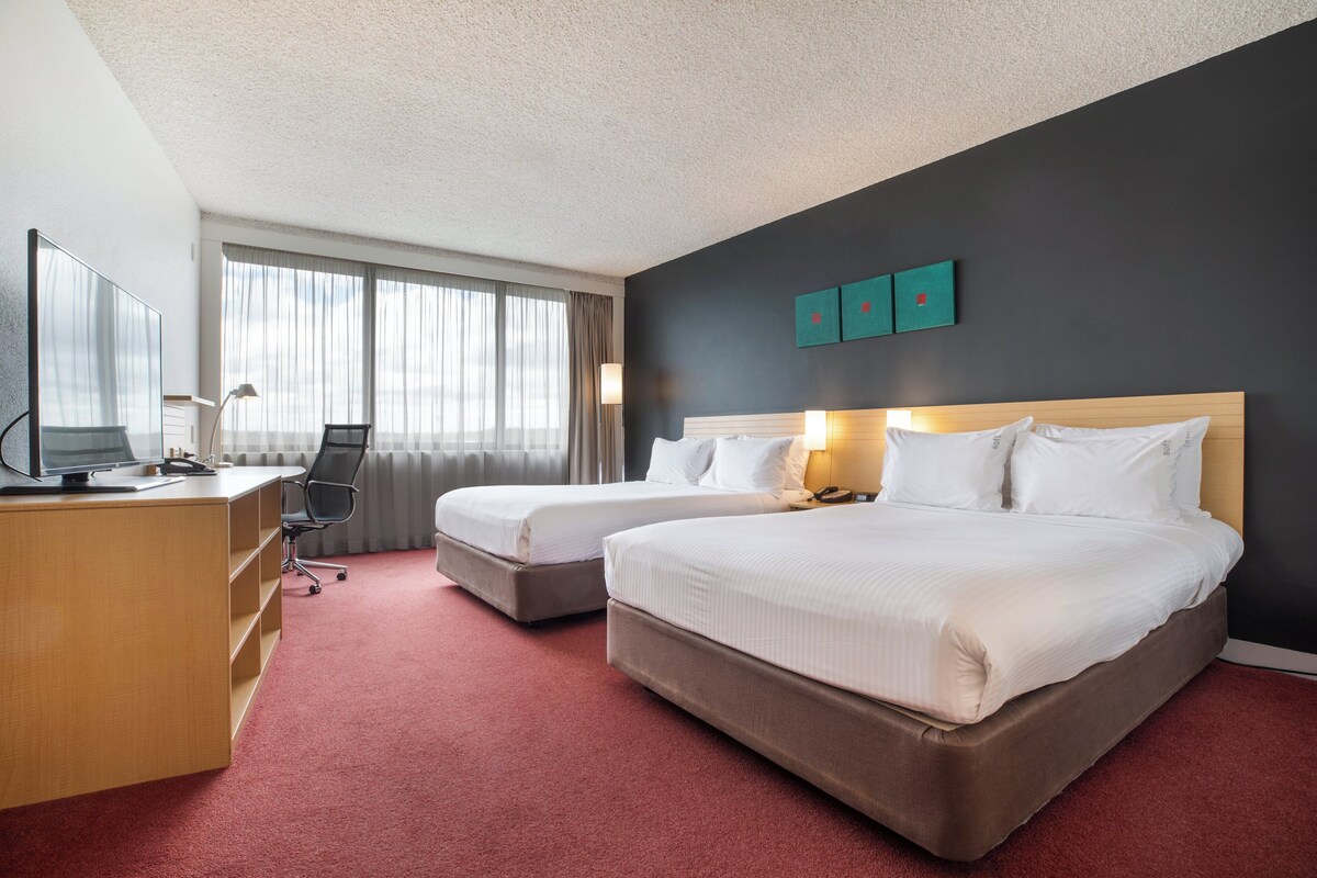 Une chambre d’hôtel avec deux lits, un bureau avec un ordinateur, une chaise et une grande fenêtre avec des rideaux.