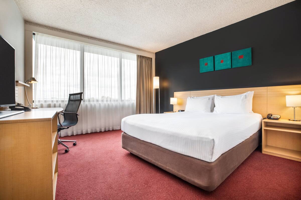 A hotel room with a large bed, a desk with a chair, a desk lamp, and a nightstand with a lamp.