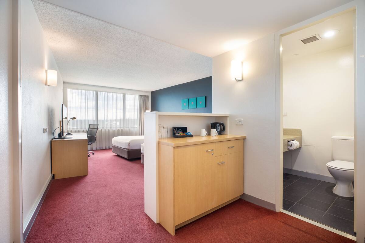 A hotel room with a bed, a desk with a chair, a wardrobe, a small bathroom with a toilet, and a window with curtains.