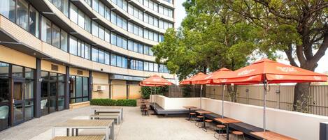 A modern outdoor seating area with tables and chairs, surrounded by a building with large windows and a tree.