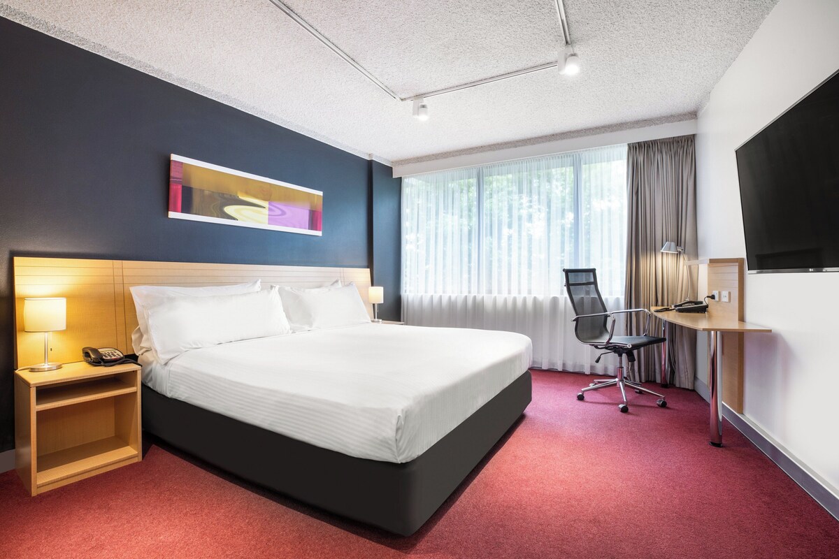 Une chambre d’hôtel avec un grand lit, un bureau avec une chaise, une télévision et une fenêtre avec des rideaux.