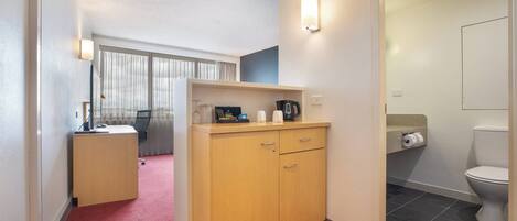 A hotel room with a wooden desk, a chair, a small cabinet, a window with curtains, a bathroom with a toilet, and a shower area.