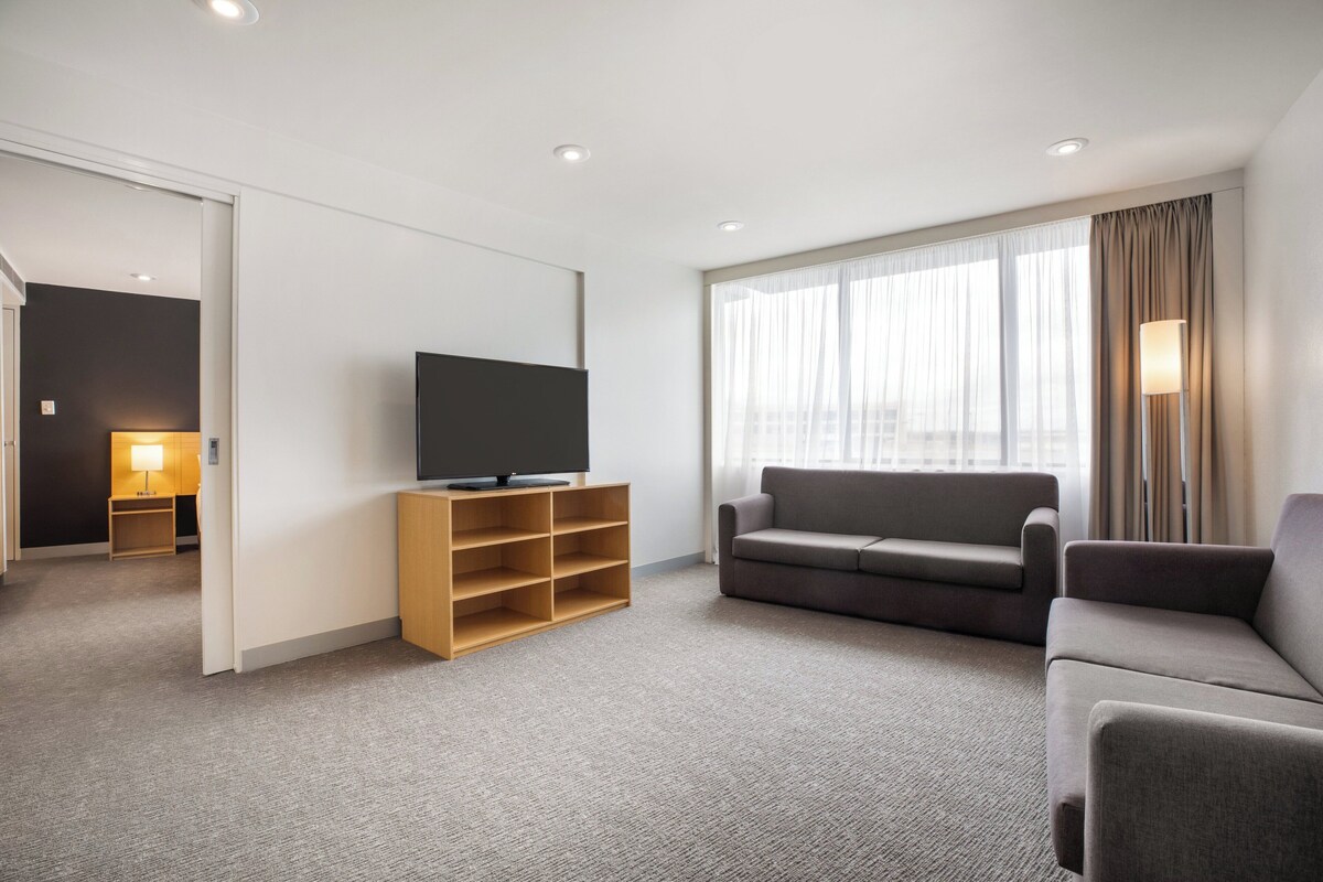 Une chambre d’hôtel moderne avec un téléviseur à écran plat posé sur un meuble en bois, un canapé gris et une grande fenêtre avec des rideaux.