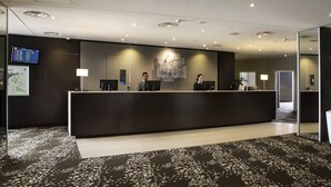 A hotel reception area with a dark reception desk, two receptionists, a digital display board, and a large potted plant.