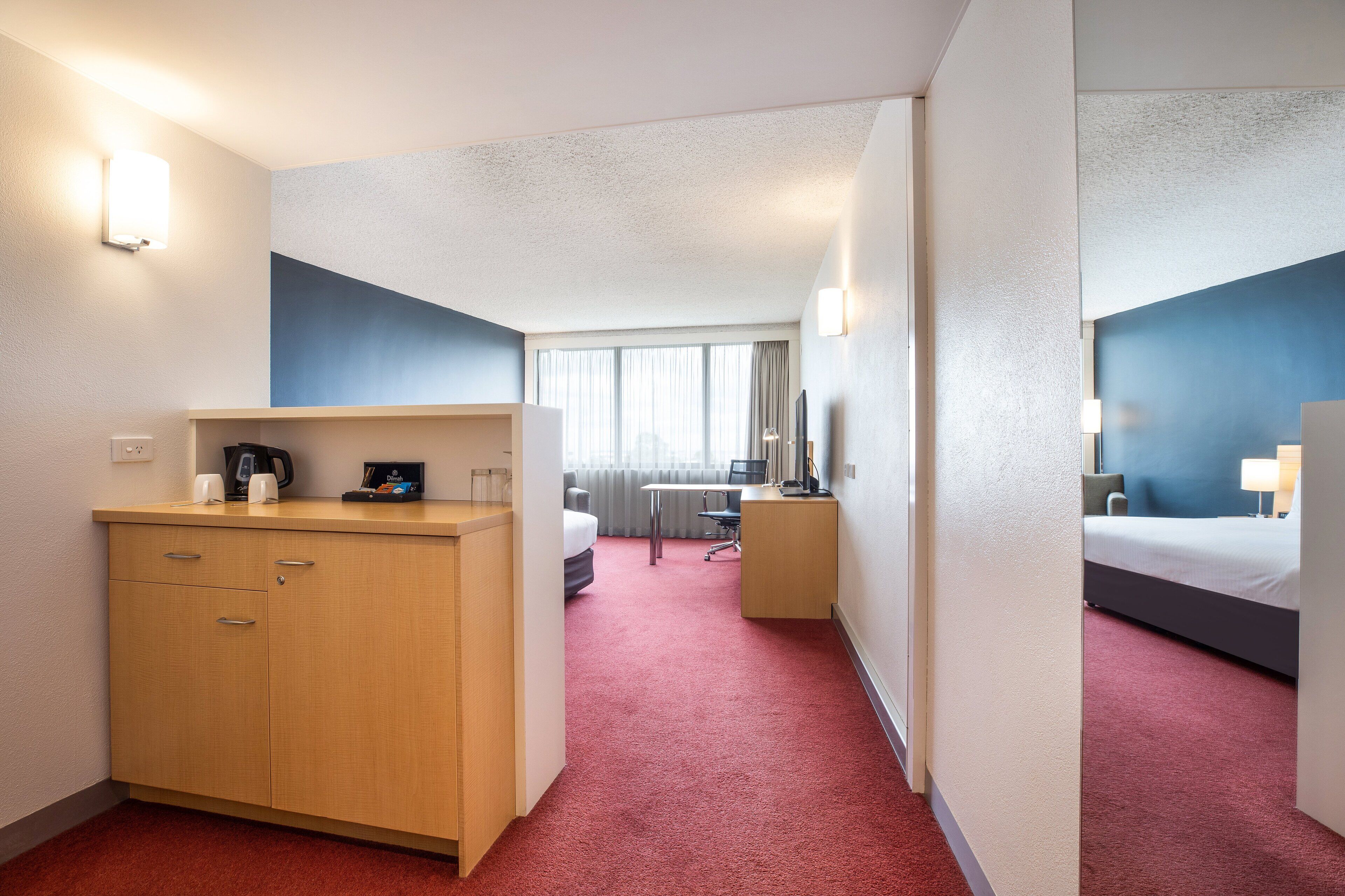 Habitación de hotel con escritorio, cama, armario y vistas a la ciudad a través de amplios ventanales.