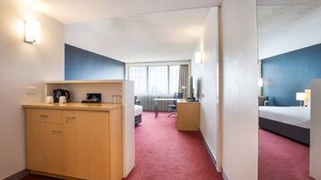 Habitación de hotel con escritorio, cama, armario y vistas a la ciudad a través de amplios ventanales.