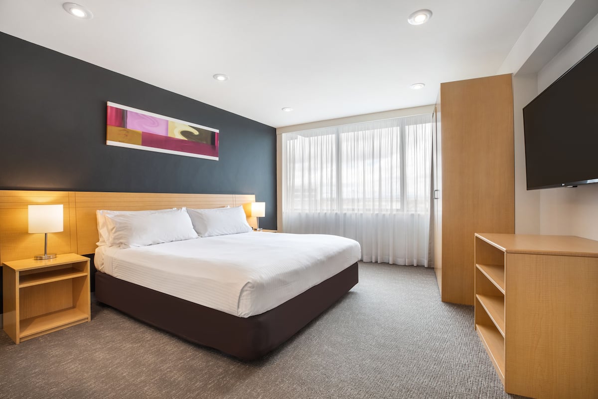 A modern hotel room with a large bed, bedside tables, a flat-screen TV, and a large window with curtains.