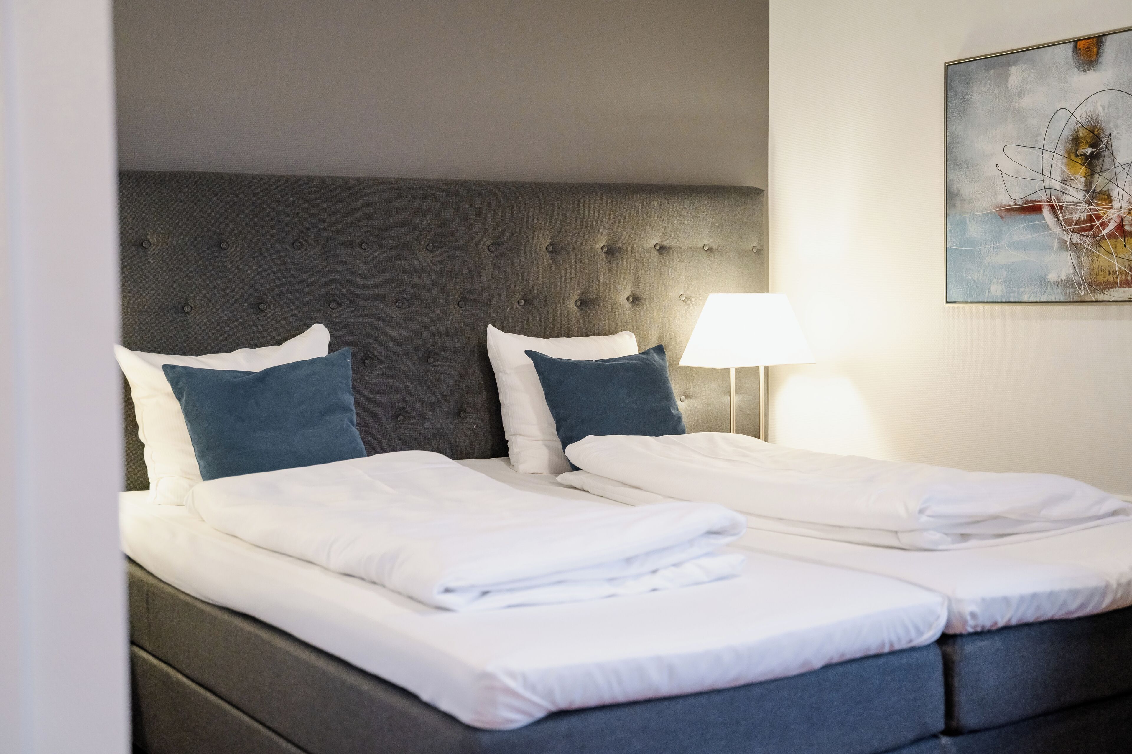 Una cama doble con cabecera capitonada, dos almohadas y una mesita de noche con lámpara.
