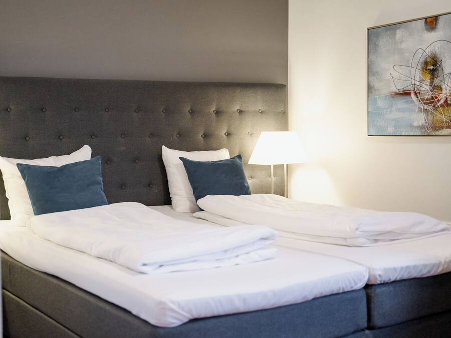 Una cama doble con cabecera capitonada, dos almohadas y una mesita de noche con lámpara.