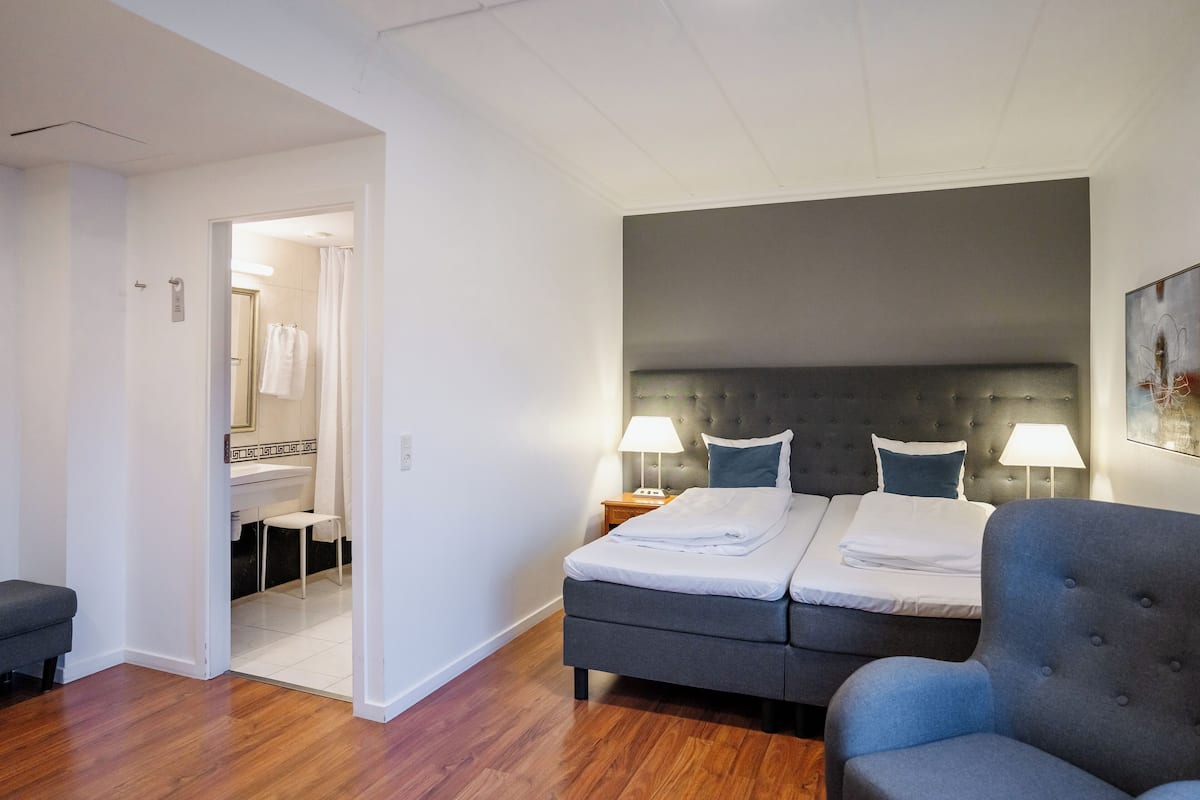 A hotel room with a double bed, two bedside tables with lamps, a grey armchair, a wooden floor, and a bathroom visible through an open door.