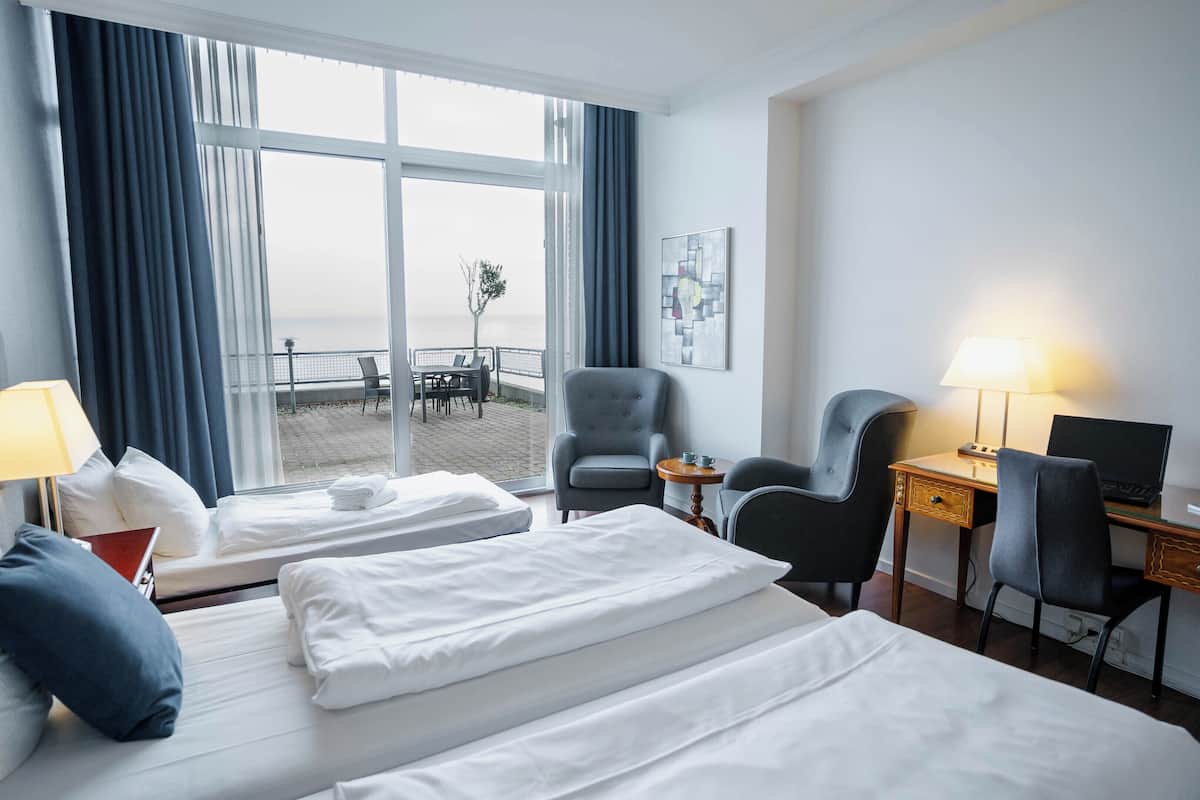 A hotel room with two beds, a desk, two chairs, a laptop, a lamp, and a view of the sea through large windows.