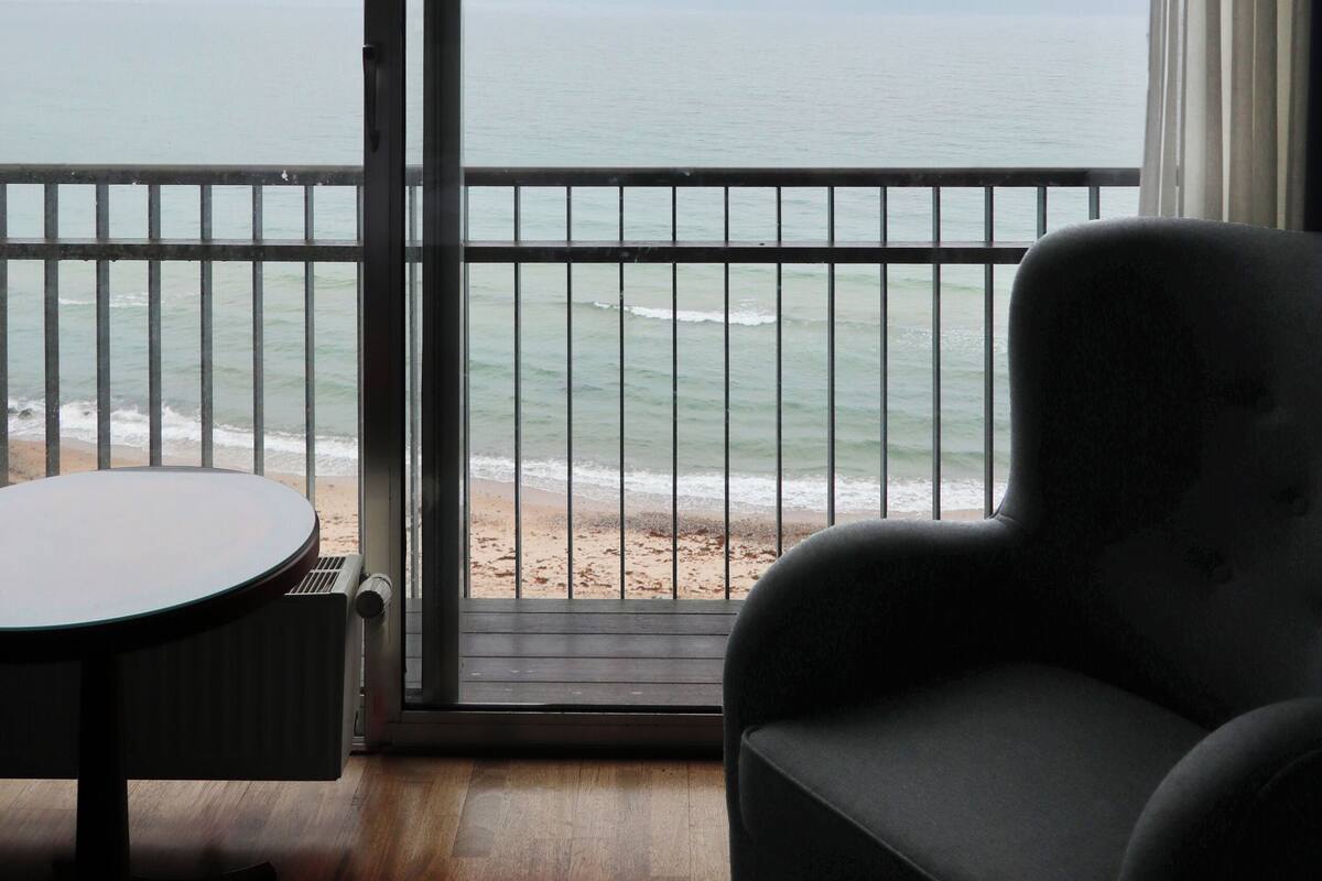 A room with a view of the ocean through a large window, a chair, and a small table.