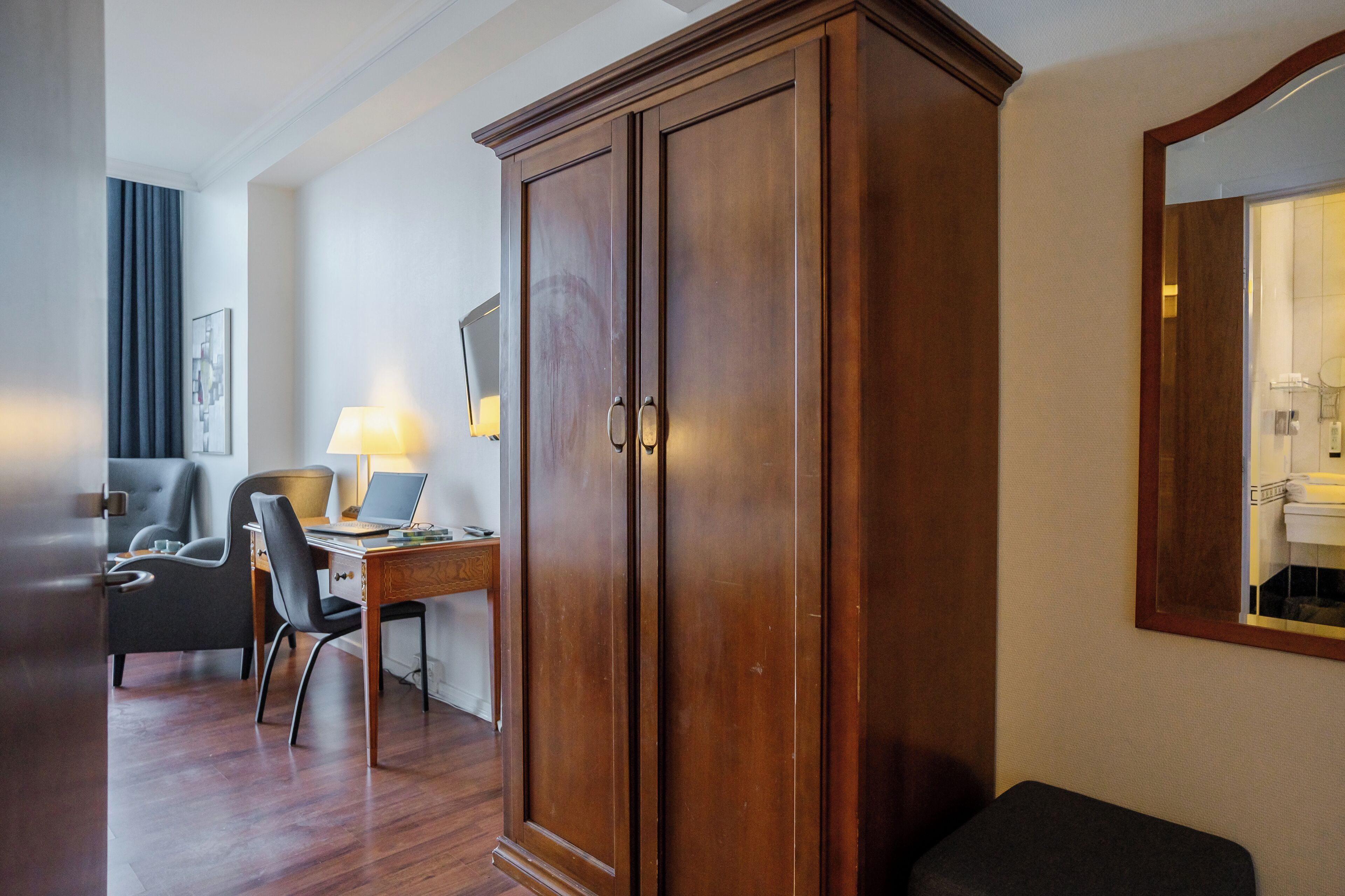 A hotel room with a wooden wardrobe, a desk with a laptop, a chair, a lamp, and a large mirror.