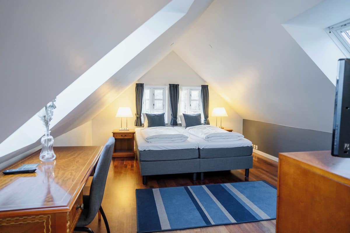 A bedroom with a sloped ceiling, a bed with pillows, a desk with a lamp, and a television on a wooden dresser.