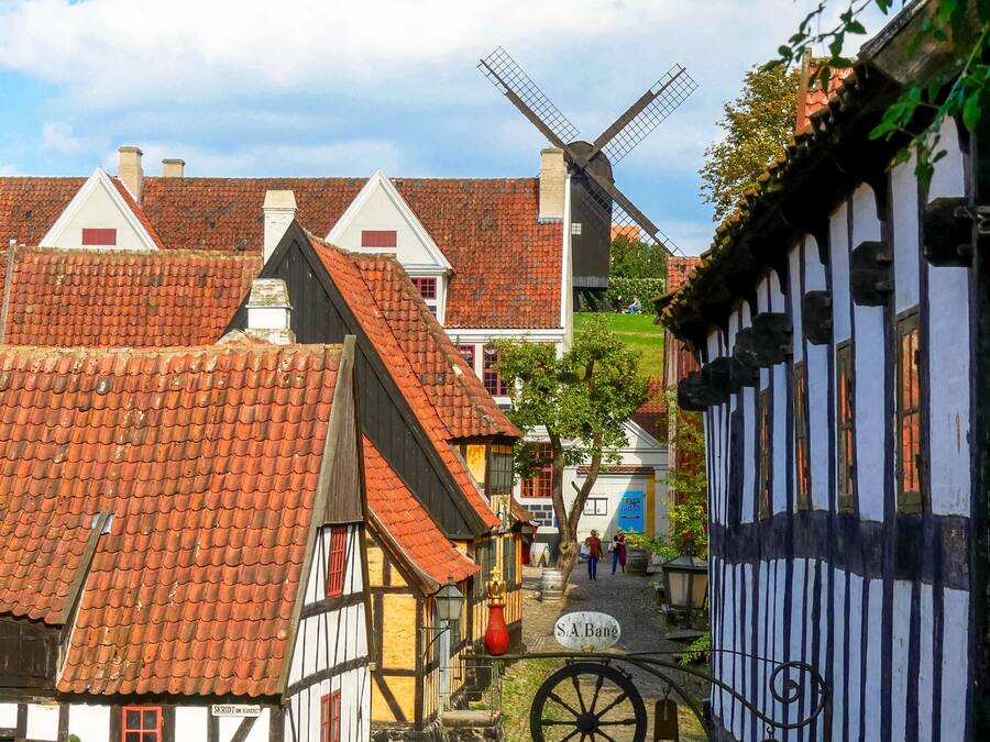 A traditional European village with timber-framed houses, a windmill, cobblestone streets, and a sign reading "S.A. Bang".
