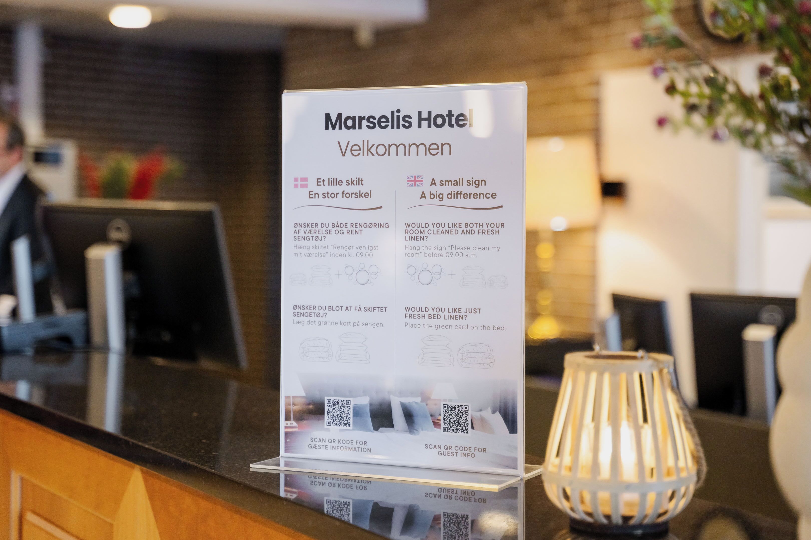 A reception desk at Marselis Hotel with a signboard providing welcome messages and QR codes for guest information.